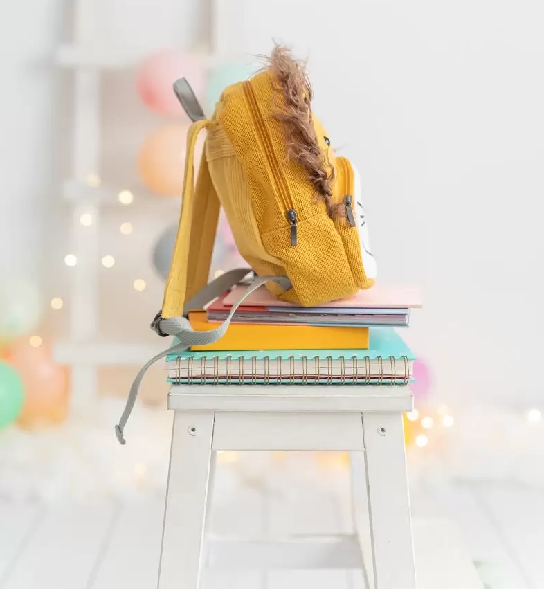 Mochila Simba Mochila infantil amarela sobre cadernos coloridos em banco branco com decoração colorida ao fundo