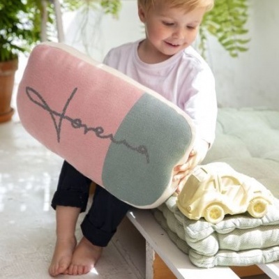 Criança segurando almofada retangular rosa e verde com texto 'Moreva', ao lado de almofadas verdes e carrinho de madeira.