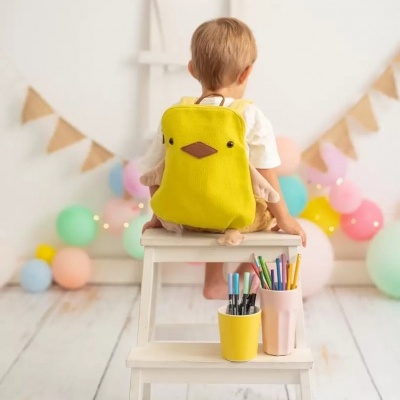 Mochila infantil amarela em forma de pato num cenário com balões e canetas