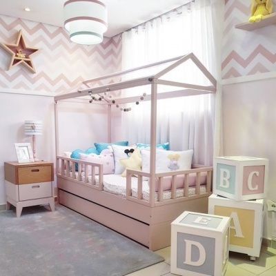 Quarto infantil com cama-casa rosa, almofadas coloridas, mesa de cabeceira e cubos decorativos com letras