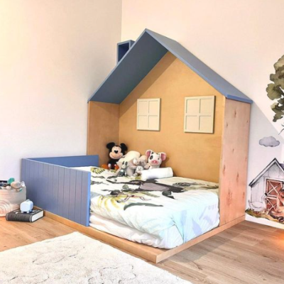 Cama infantil em forma de casa com roupa de cama e peluches num quarto com piso em madeira e tapete branco.