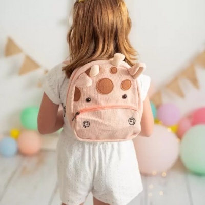Criança com mochila rosa em forma de girafa, cenário claro e festivo ao fundo