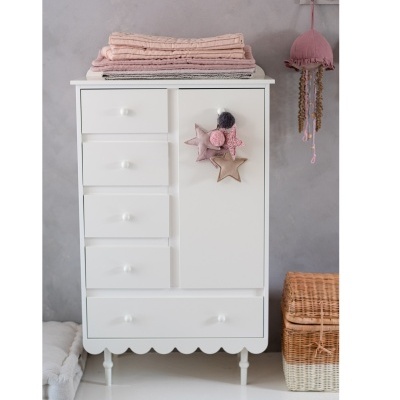 Móvel branco com gavetas e porta decorada, mantas cor-de-rosa em cima, cesto de verga e pendente rosa ao lado.