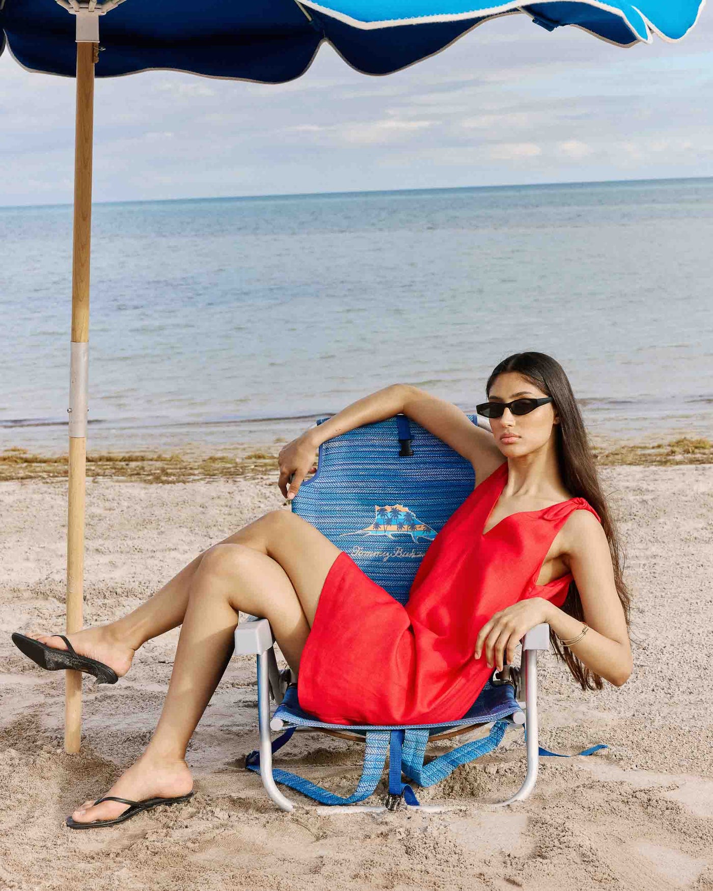 Wynette Linen Blend Dress Mulher sentada numa cadeira de praia azul, vestindo vestido vermelho e óculos de sol pretos, na areia junto ao mar