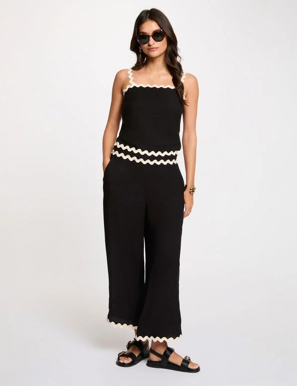 Popa Cropped Trousers Mulher com macacão preto de alças com detalhes creme, sandálias pretas e óculos escuros em fundo branco.