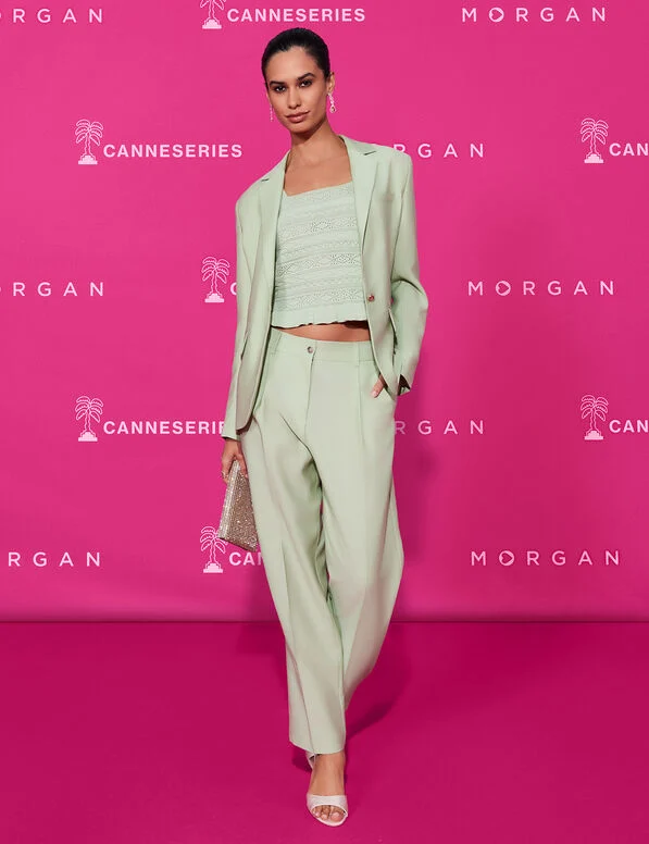 Medi Carrot Trousers Modelo veste conjunto verde claro com blazer, calças e top de renda, segura mala brilhante, em fundo rosa com textos.