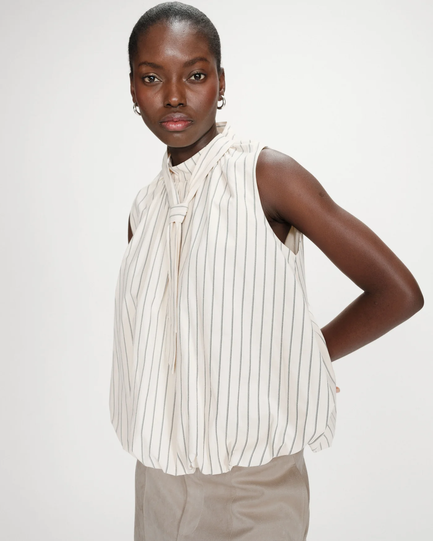 Vanya Blouse Blusa branca às riscas verticais pretas com nó ao pescoço, usada por mulher em fundo branco.