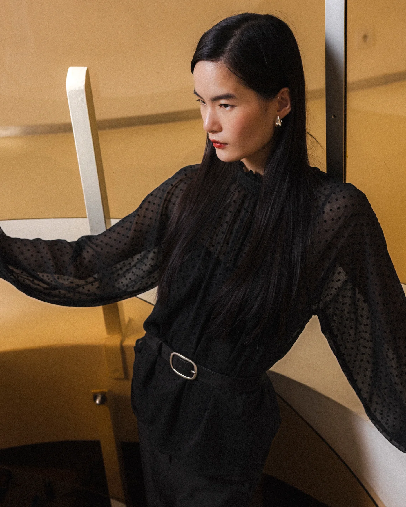 Tailly Blouse Blusa preta transparente de bolinhas com cinto preto em modelo junto a escadas interiores.