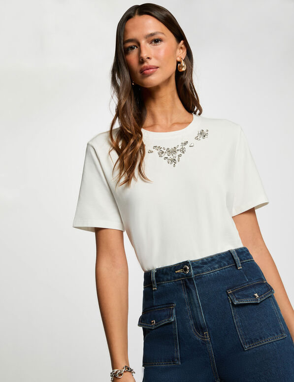 mare t-shirt Mulher a usar t-shirt branca com bordado floral prateado no decote e saia jeans azul escura com bolsos frontais