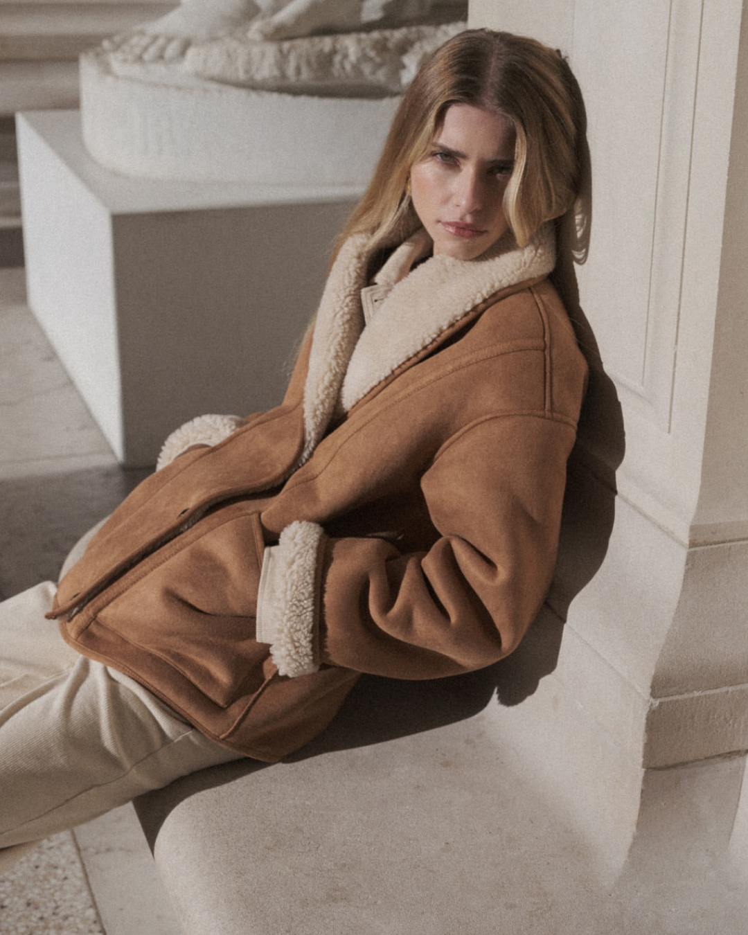 swan coat Mulher com casaco castanho de veludo com forro e gola de pelo branco creme sentada junto a uma parede