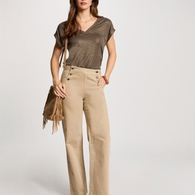 Mulher com t-shirt castanha metálica, calças cáqui com botões na cintura, botas castanhas e bolsa com franjas