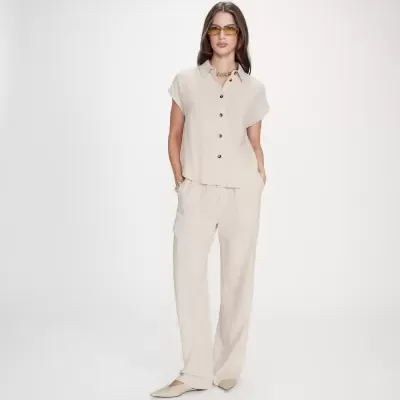 Mulher vestindo conjunto bege claro com camisa de manga curta e calças compridas, sapatos baixos bege e acessórios dourados em fundo branco.