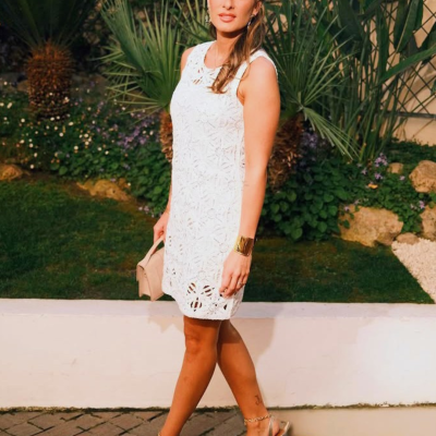 Mulher com vestido branco de renda e sandálias douradas num jardim.