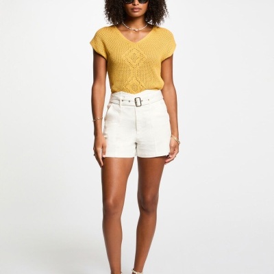 Mulher com camiseta amarela de malha, calções brancos com cinto, sandálias bege com tachas, e óculos escuros