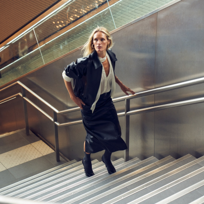 Mulher a subir escadas com casaco preto, camisa branca e saia preta midi