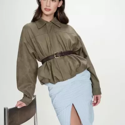 Mulher com casaco verde-oliva, saia azul clara riscada, cinto castanho e cadeira