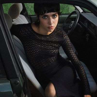 Mulher sentada no banco dianteiro de um carro com blusa preta transparente e saia preta.