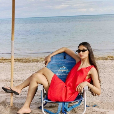 Mulher sentada numa cadeira de praia azul, vestindo vestido vermelho e óculos de sol pretos, na areia junto ao mar