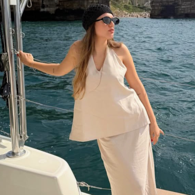Mulher com vestido bege longo e boina preta em barco com mar ao fundo