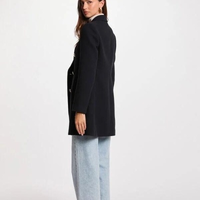 Pessoa com casaco preto, calças de ganga claras e sapatilhas brancas e azuis, fundo branco