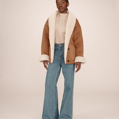 Mulher com casaco bege de pelo branco, camisola bege e jeans azuis largos em fundo claro