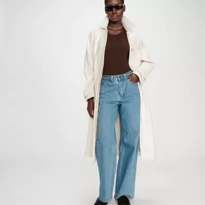 Pessoa com casaco comprido branco, camisa castanha, jeans azuis e sapatos pretos.