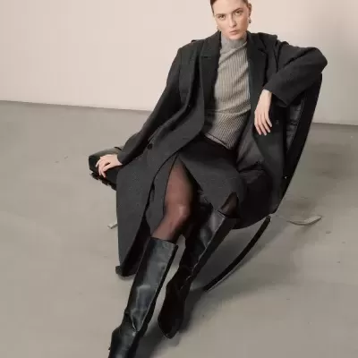 Mulher sentada de casaco cinzento escuro, saia, camisa cinza de gola alta, botas pretas e collants pretos em cadeira preta.
