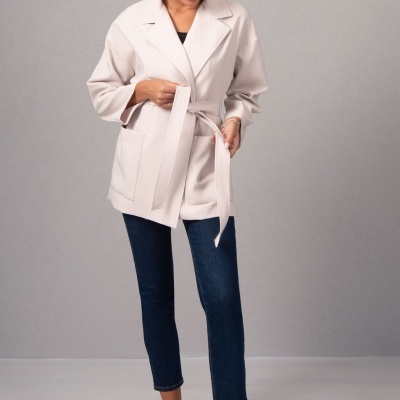 Casaco bege claro com cinto e gola de lapela usado por mulher com calças de ganga e sapatos pretos
