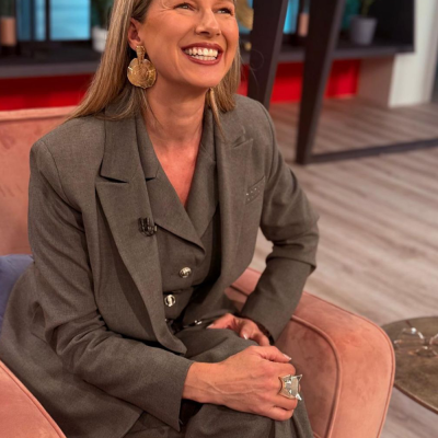 Mulher sentada com fato cinzento, brincos grandes dourados e anel prateado, em cadeira rosa no interior.