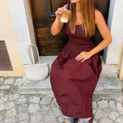 Mulher sentada numa soleira junto a uma porta de madeira, vestindo vestido bordeaux, sandálias estampadas azuis e bordô, com bolsa branca trançada ao lado e bebida em copo plástico.