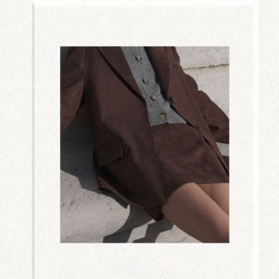 Blazer e saia castanhos com camisola cinzenta e botões, sentada junto a uma parede clara.