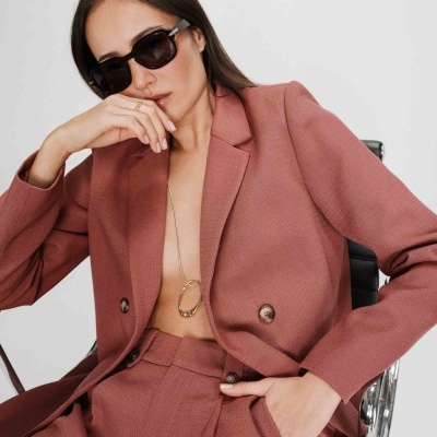Mulher com fato cor tijolo, óculos de sol e joias douradas, sentada em cadeira