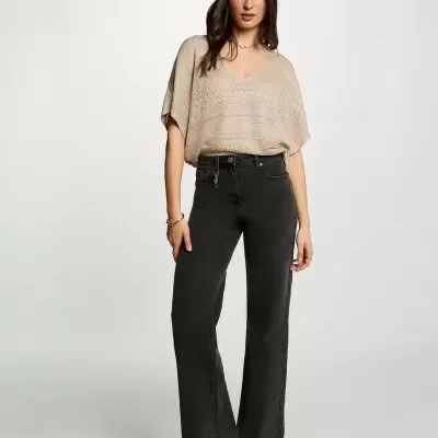 Mulher com camisola bege de malha, calças pretas de ganga e sandálias castanhas em fundo cinzento