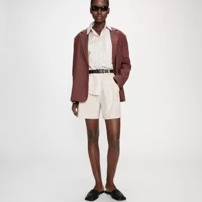 Mulher de blazer castanho, camisa creme riscada, calções bege, mules pretos e óculos escuros