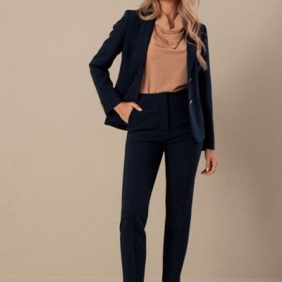 Mulher vestindo blazer e calças azul escuro, blusa castanha clara, e sapatos de salto bege em fundo cinza neutro.