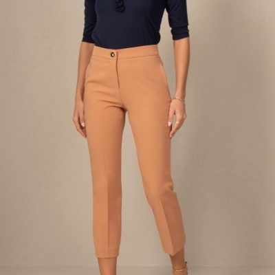 Mulher com blusa azul escura de babados e calças bege claras, sandálias douradas, em fundo cinza claro