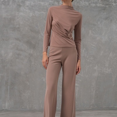 Mulher com conjunto castanho claro de blusa com fivela dourada e calças largas, sapatos bege, fundo cinzento.