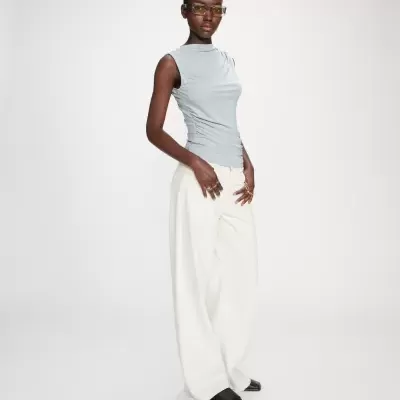 Modelo com top cinza claro, calças brancas largas, sapatos pretos e óculos amarelos em fundo branco