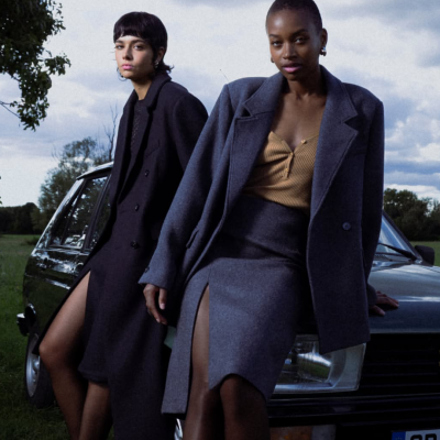 Duas mulheres com casacos compridos e fendas laterais apoiadas num carro antigo preto ao ar livre.