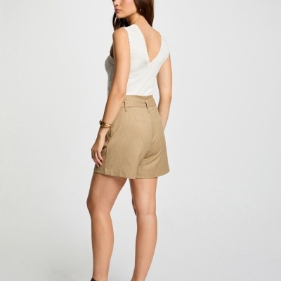 Mulher a usar blusa branca sem mangas e shorts bege com sapatos pretos num fundo branco