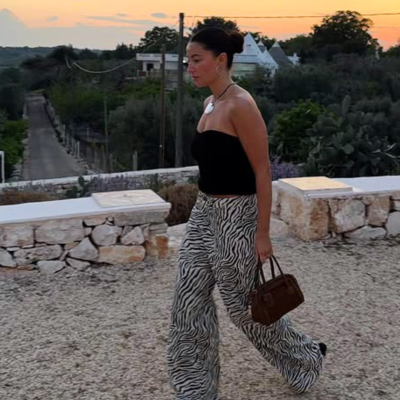 Mulher com top preto e calças zebrada a caminhar ao pôr do sol segurando mala castanha