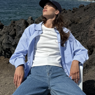 Mulher sentada ao ar livre com camisa azul às riscas, t-shirt branca, jeans e boné preto.