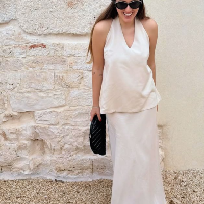 Mulher com roupa creme e acessórios pretos, parede de pedra clara ao fundo.