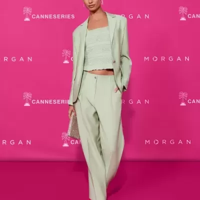 Modelo veste conjunto verde claro com blazer, calças e top de renda, segura mala brilhante, em fundo rosa com textos.