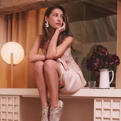 Mulher com vestido rosa claro e sapatilhas bege com detalhes dourados, sentada numa bancada branca em ambiente interior com madeira e cortinas.