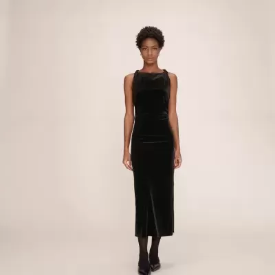 Vestido longo preto de veludo com alças ajustadas, usado por mulher com meias e sapatos pretos, fundo neutro.