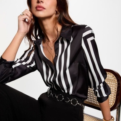 Mulher sentada com camisa preta e branca e calças pretas numa cadeira com vime