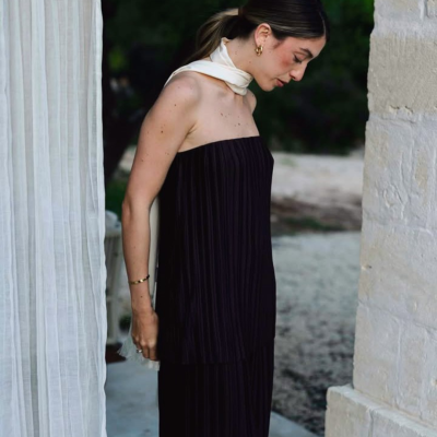 Mulher vestindo conjunto preto plissado e lenço branco ao pescoço em ambiente externo com parede de pedra e cortina branca.