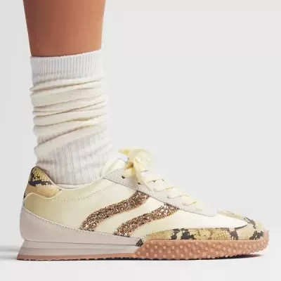 Ténis creme com detalhes em pele de cobra e brilho, solo de borracha texturada, usado com meias brancas.
