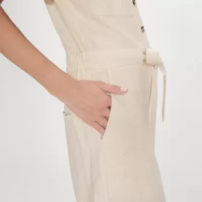 Vestido bege de tecido natural com bolsos e cinto, vista lateral com mão no bolso, fundo branco.